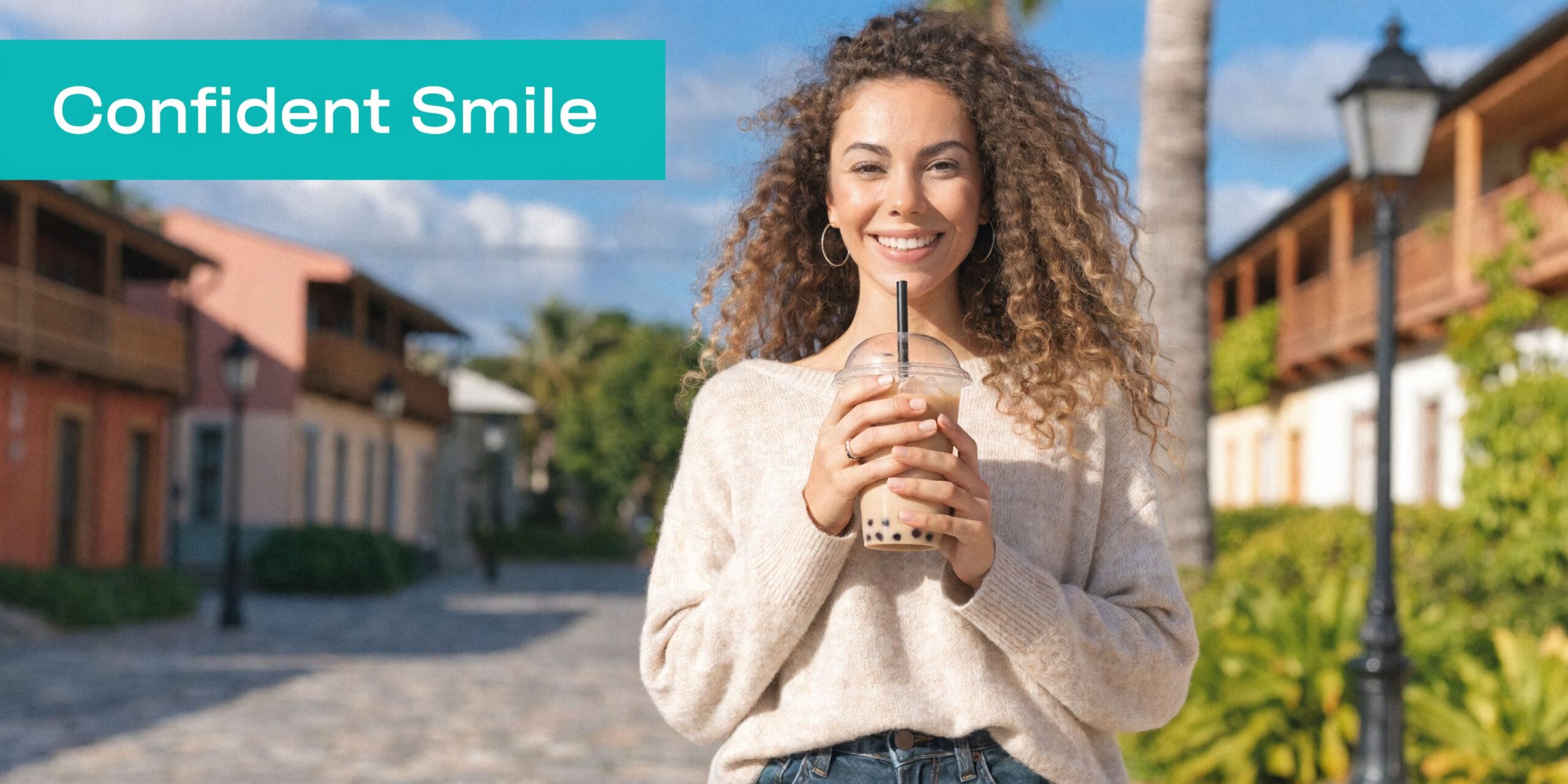 A happy woman with curly hair smiles confidently while holding a refreshing cup of bubble tea outdoors.