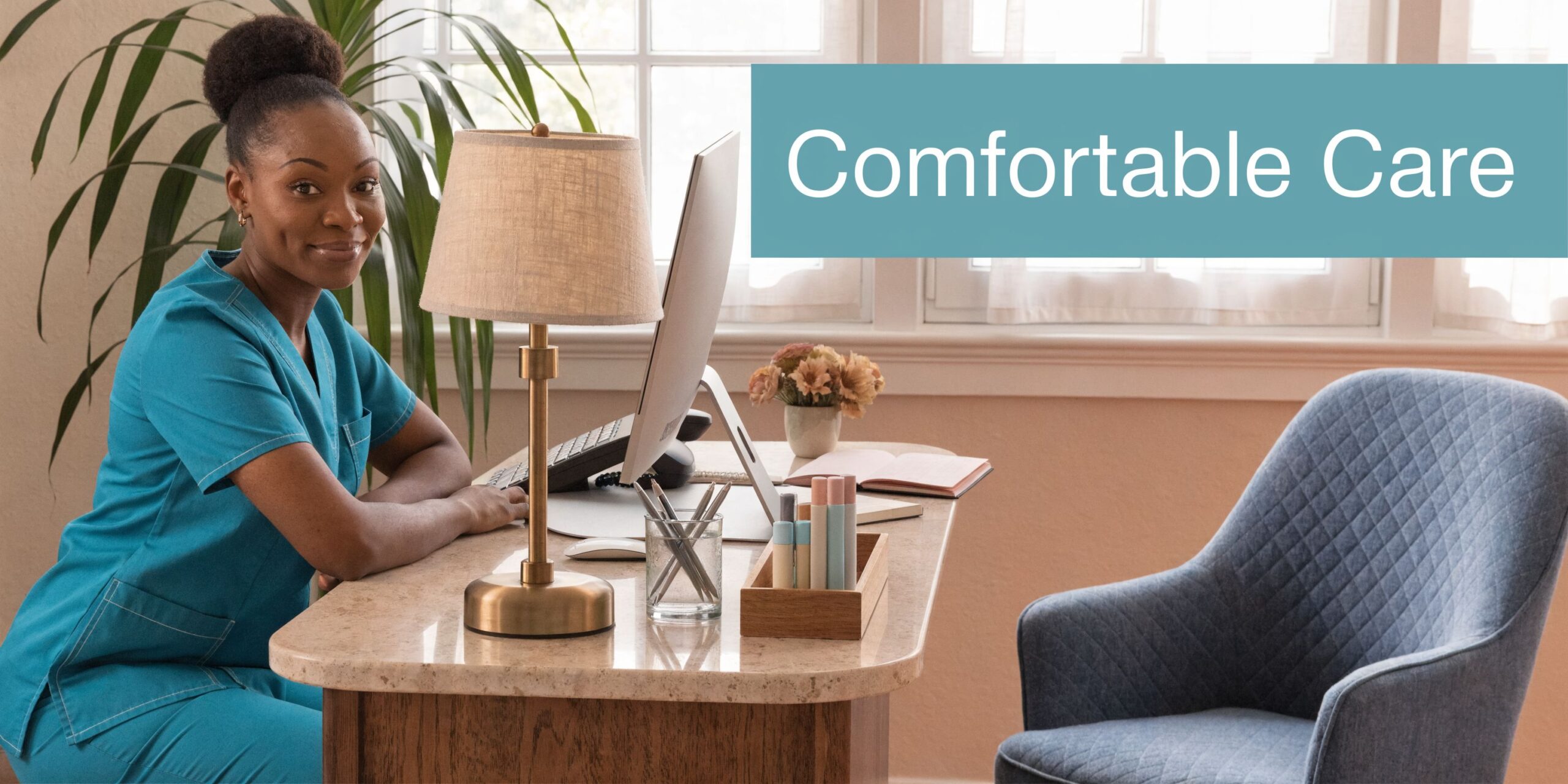 A friendly healthcare professional in blue scrubs sitting at a clean, well-organized office desk with a lamp.