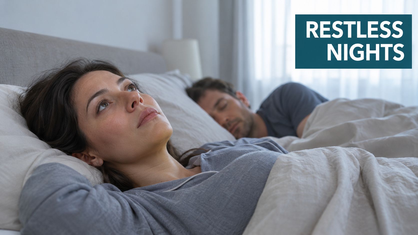 A woman lying awake in bed at night looking stressed while her partner sleeps peacefully nearby.